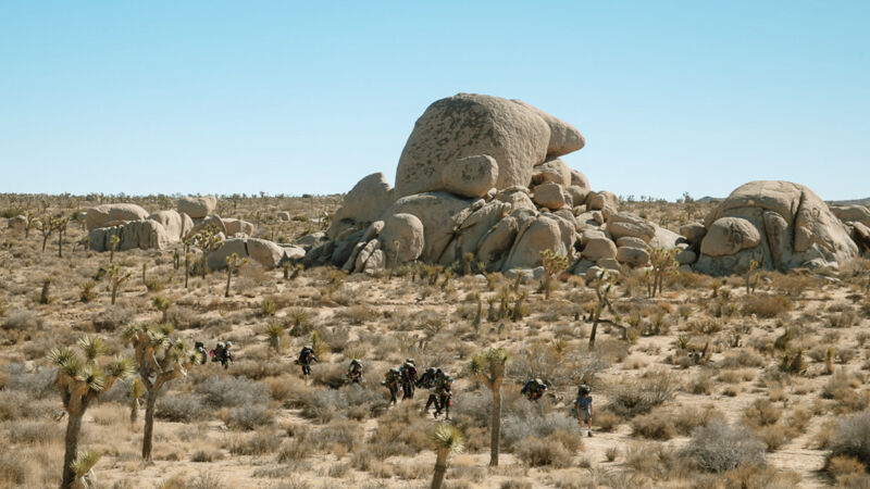 The image shows a desert landscape with sparse vegetation and large rock formations. A group of people are walking across the desert floor, appearing small in the vastness of the scene. The sky is clear and blue, suggesting a sunny day. The overall impression is one of a remote and arid environment.